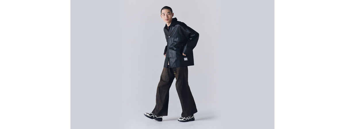 Model wearing black leather jacket, wide dark brown trousers, and black and white Asics Gel-1130 sneakers, standing against a plain grey background.