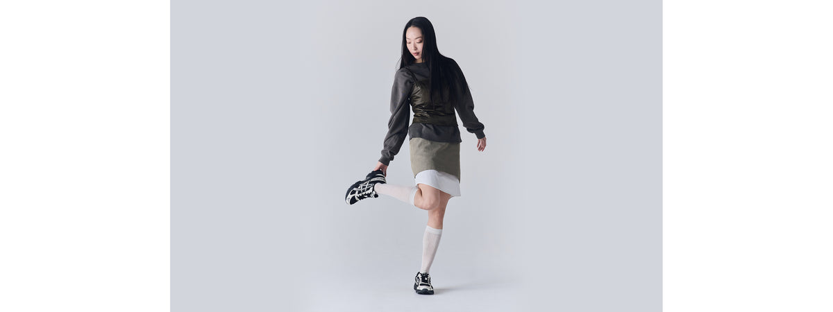 A woman wearing a dark sweatshirt and skirt with white knee-high socks poses lifting one leg, showing black and white Asics Gel-1130 sneakers on both feet.