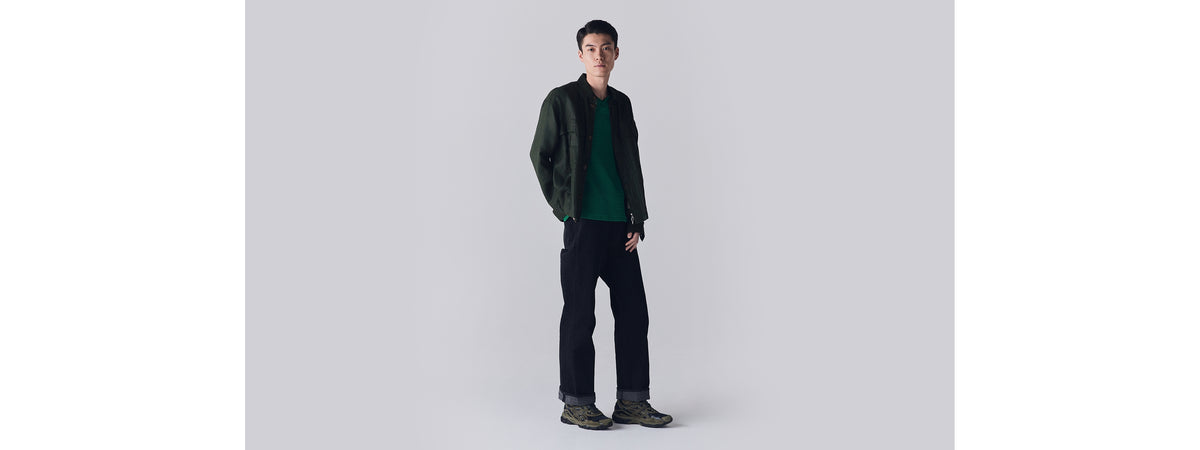 Man standing in green jacket, green sweater, and black jeans wearing green and gray Asics Gel-NYC sneakers.