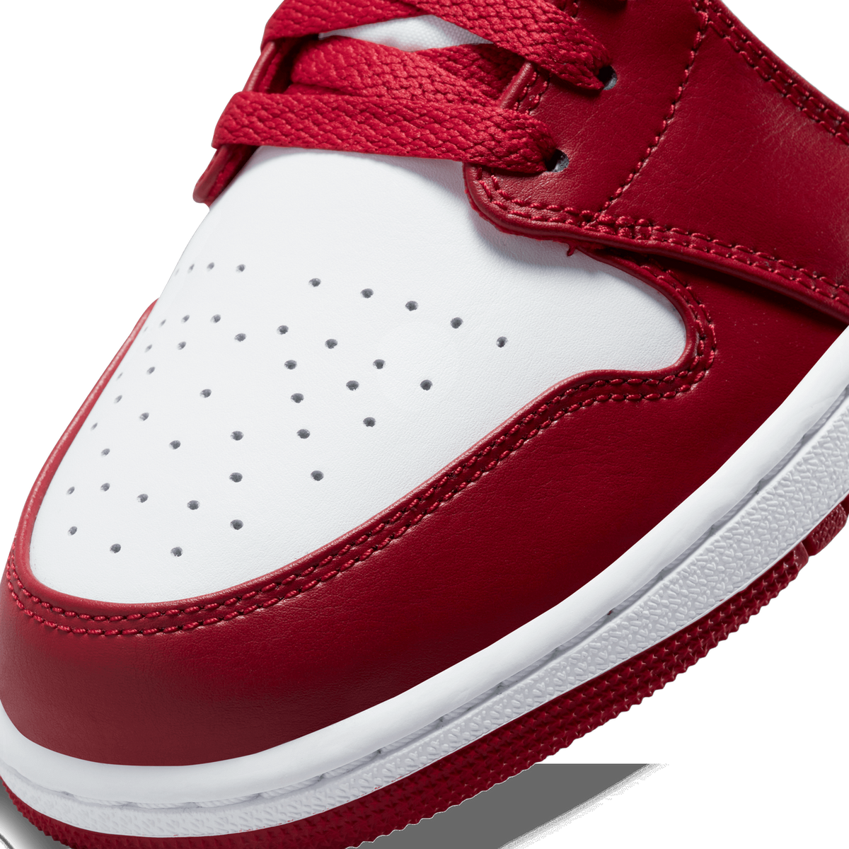 Extreme close-up of the toe box and lacing of a red and white Air Jordan 1 Low, showing perforated white leather on the toe, red leather overlays with stitched detailing, red laces, and the white midsole edge with red rubber outsole.