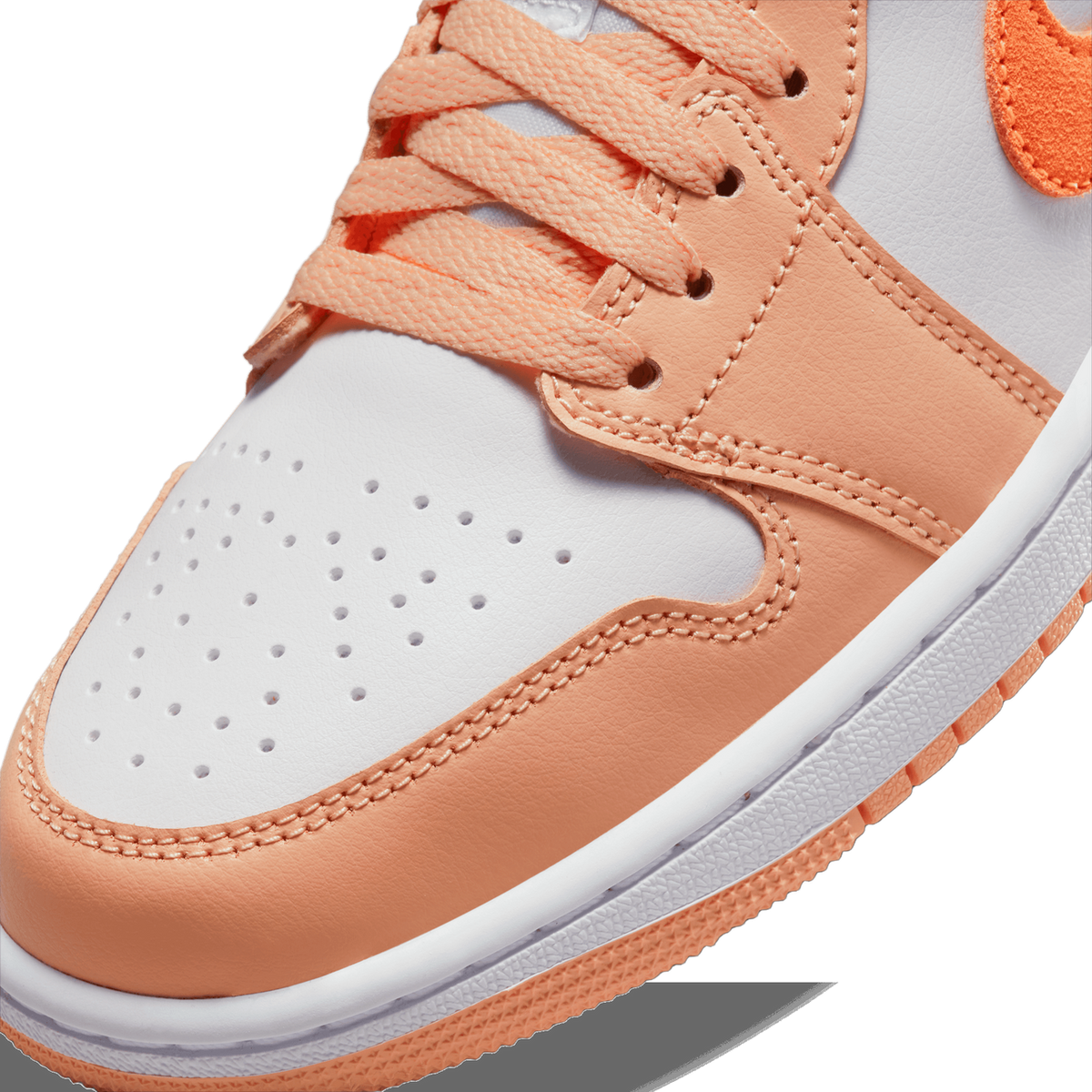 Close-up of the toe box and lacing of a peach-orange and white Air Jordan 1 Low, showing perforated white toe leather, orange overlays with contrast stitching, peach laces, and an orange Swoosh logo on the upper.