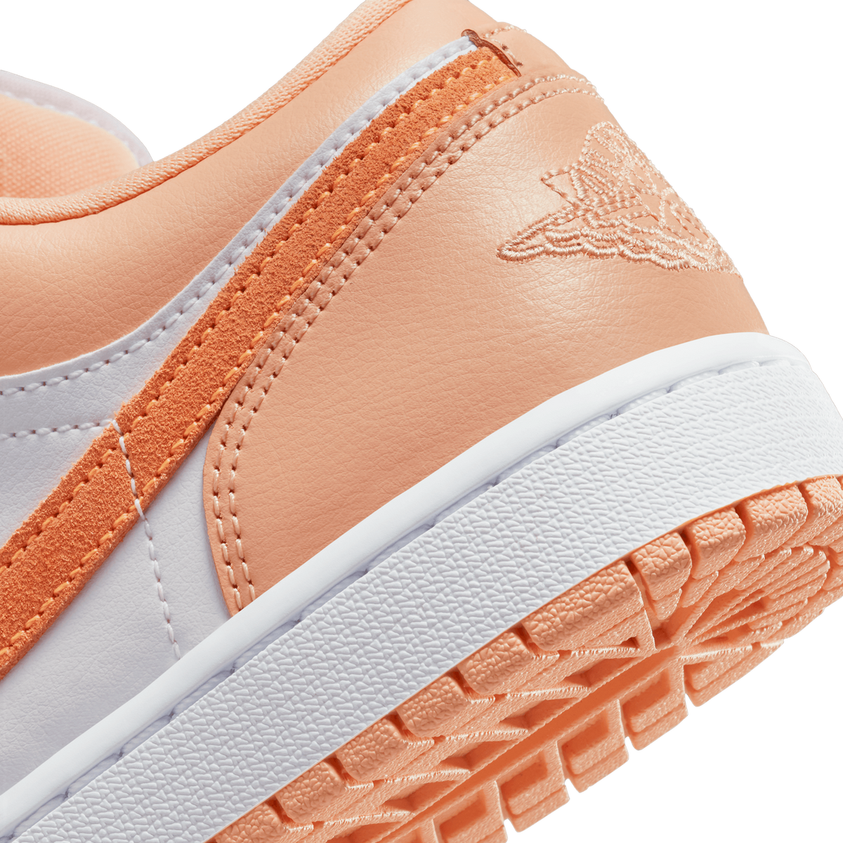 Close-up of the heel and lower side of a peach-orange and white Air Jordan 1 Low, showing the embroidered Jumpman wings logo on the heel, orange suede overlay stripe, and the white midsole with peach-coloured rubber outsole lugs.