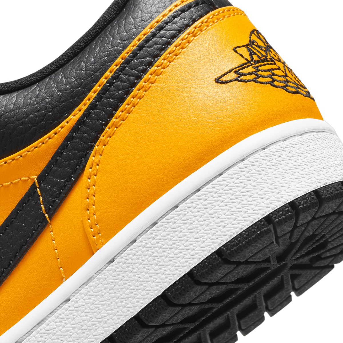 Extreme close-up of the heel and rear quarter of a gold, black and white low-top sneaker, showing the embroidered black Jordan Wings logo on the gold heel, black pebbled leather panel, and white midsole with black rubber outsole tread.