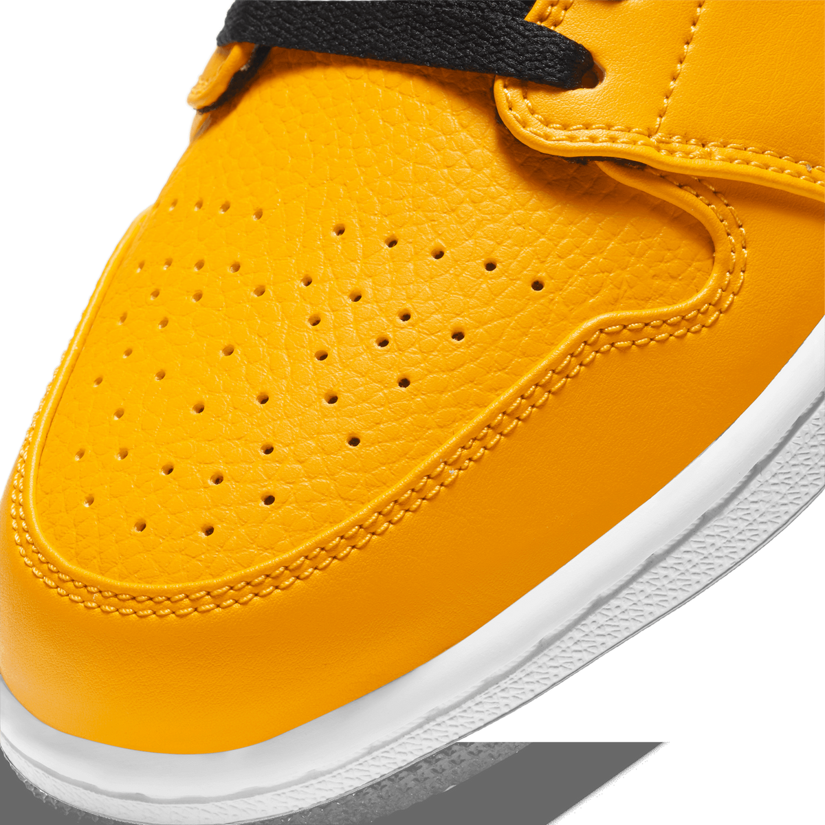 Close-up detail of the toe box of a yellow Air Jordan 1 Low, showing perforated and textured yellow leather, black laces at the top, and a white midsole with black outsole edge visible at the bottom right.