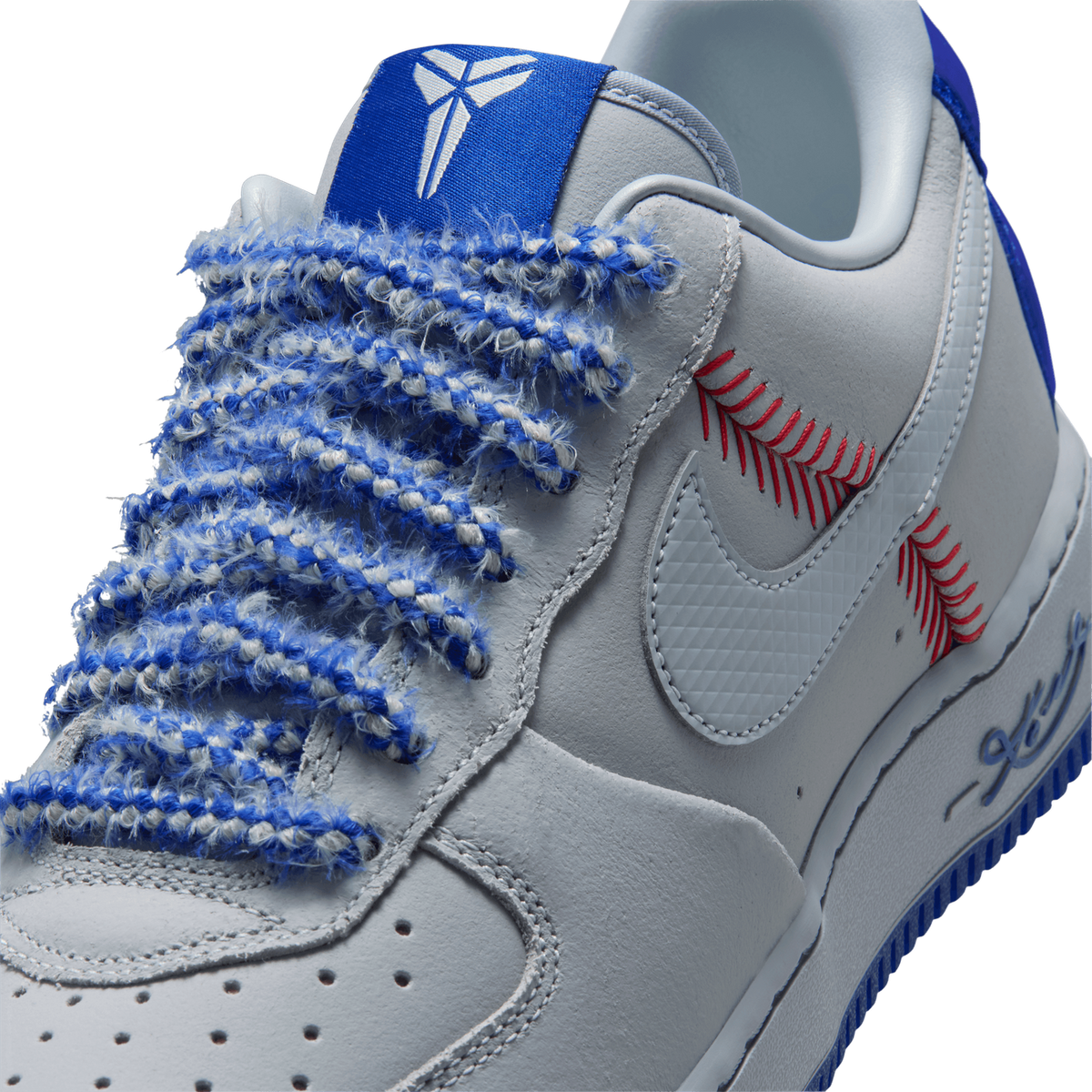 Close-up of a white and grey sneaker toe box with blue frayed laces, a Kobe Bryant mamba logo on the blue tongue, red baseball-stitch embroidery, and a grey Nike Swoosh on the lateral side.