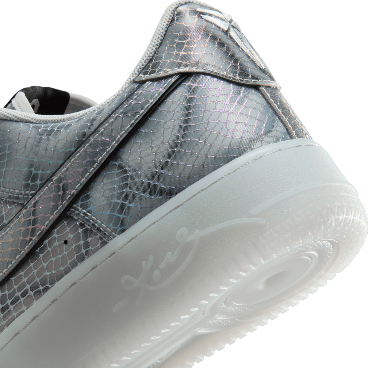Close-up of the heel and outer side of a silver, iridescent snake-scale patterned sneaker with black swoosh overlay, a translucent frosted white sole, and a script signature visible on the midsole.