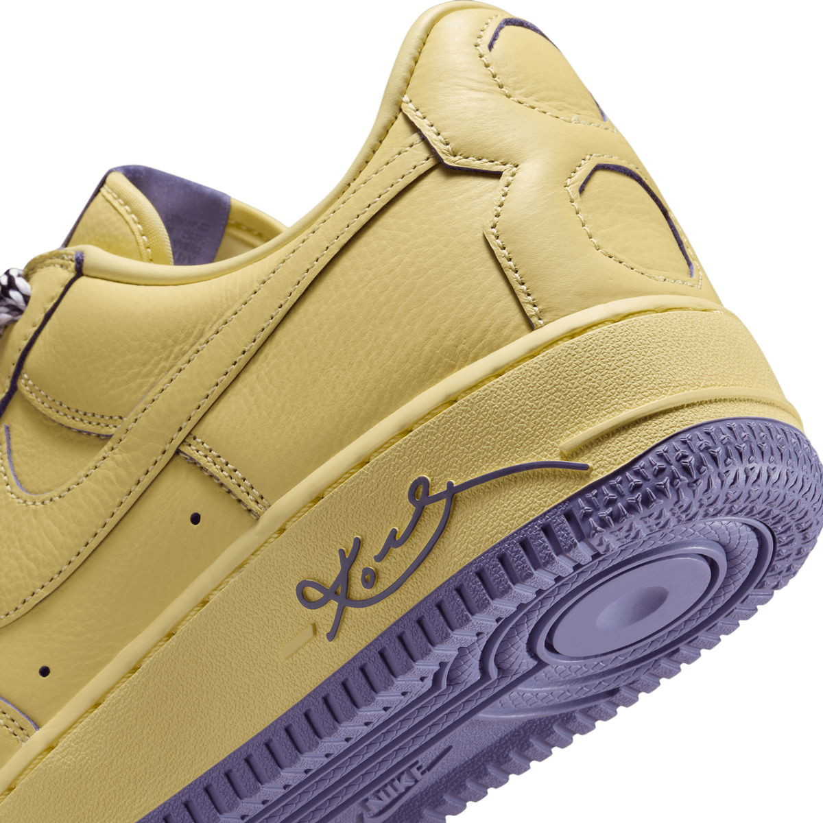 Close-up of the rear heel and outer side of a yellow leather sneaker with purple accents, showing the Kobe signature script on the midsole and a purple, tread-patterned outsole.