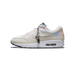 Side view of a white and cream Nike Air Max 1 sneaker with iridescent overlays, a navy heel tab, and a silver charm bracelet with a '3.26' tag hanging from the laces.