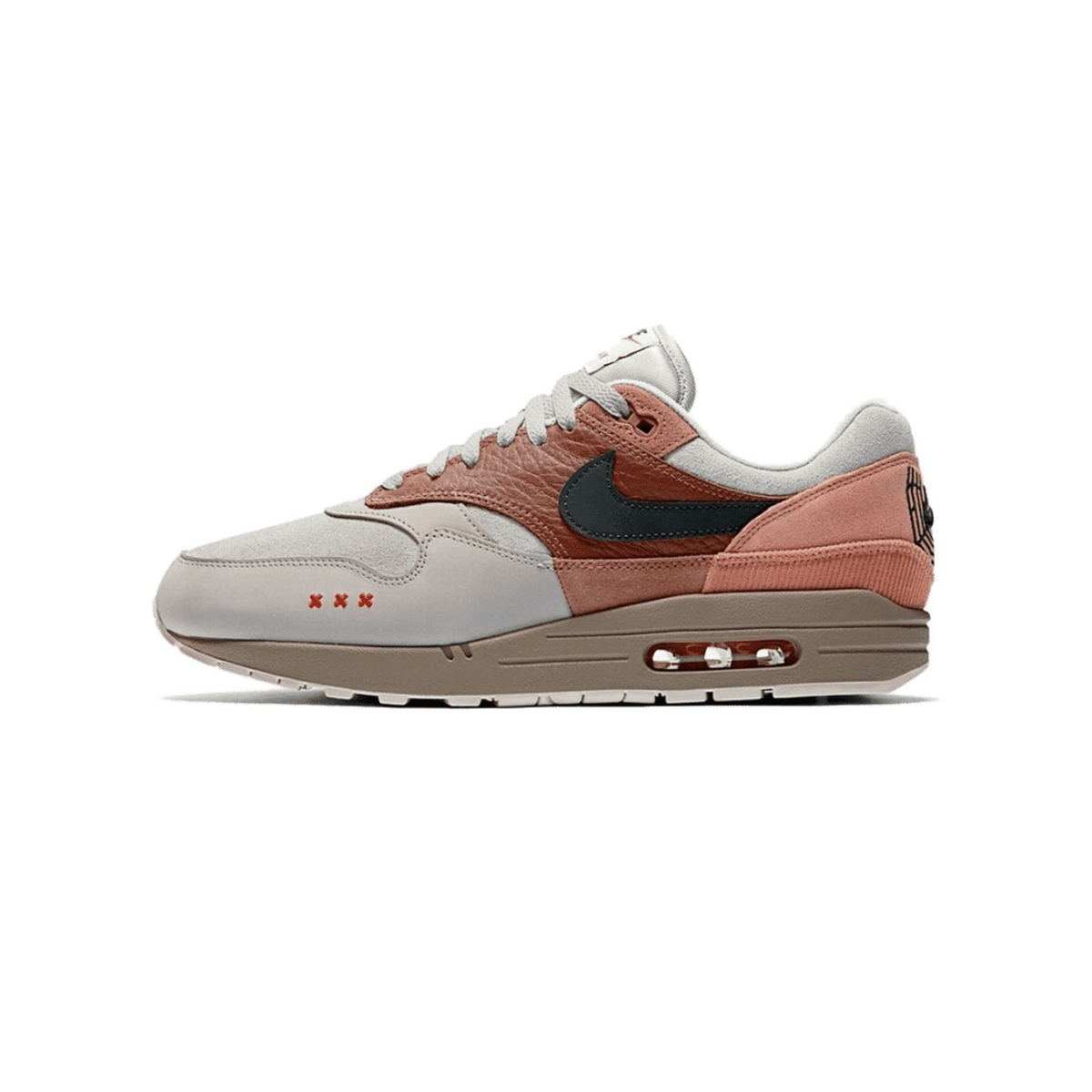 Side view of a grey and terracotta Nike Air Max 1 sneaker with a dark swoosh, corduroy heel panel, and small orange X marks on the toe box, toe pointing left.