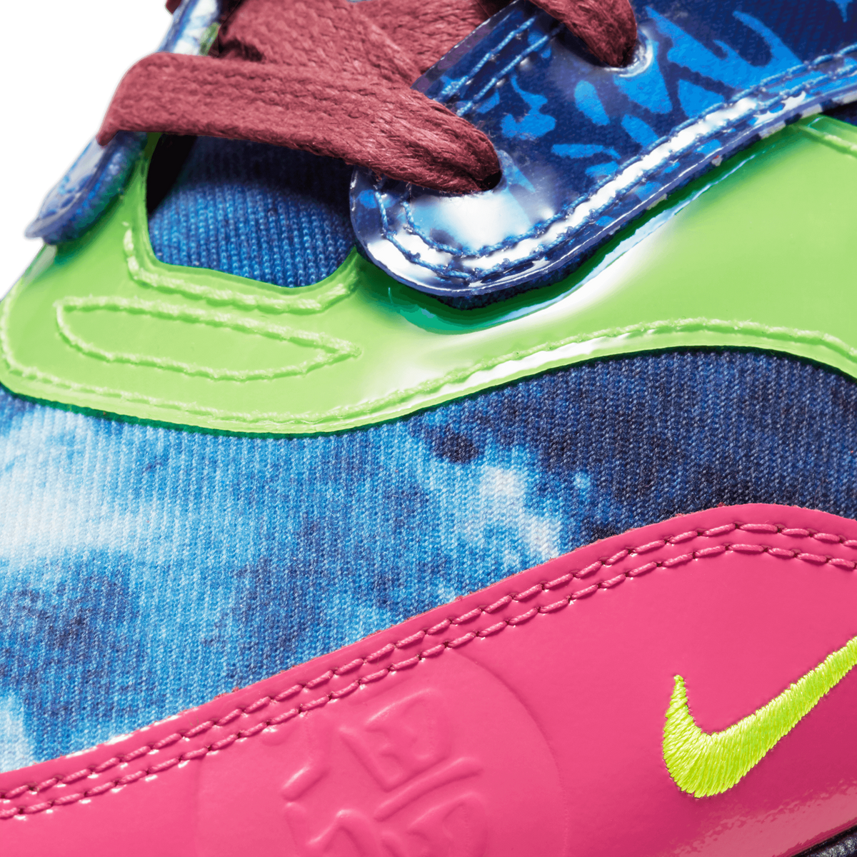Extreme close-up of a Nike Air Max 1 showing maroon laces, blue tie-dye patterned overlay, a neon green eyelet panel, and a pink embroidered Nike swoosh in volt green.