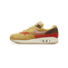 Left Nike Air Max 1 sneaker in wheat gold with rust red corduroy panel, pink fuzzy swoosh, and brown suede heel, shown from the outside with toe pointing left.