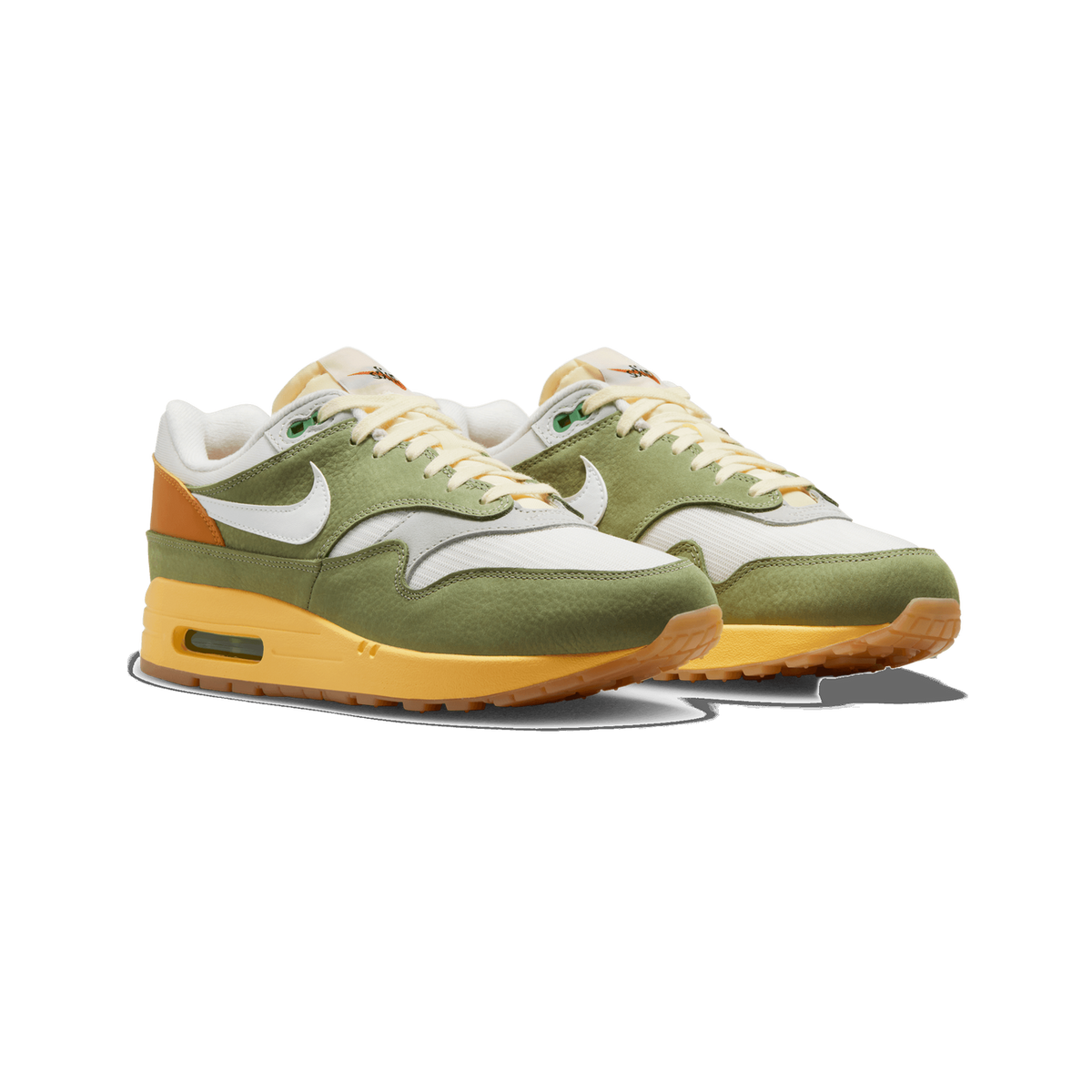 A pair of green, cream and yellow Nike Air Max 1 sneakers shown side by side at a three-quarter angle, with olive-green nubuck overlays, a white Swoosh and a golden-yellow Air Max sole.