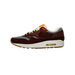 Left Nike Air Max 1 sneaker in dark brown suede with houndstooth panels, orange tongue tab, cream laces and midsole, shown from the side with toe pointing left.