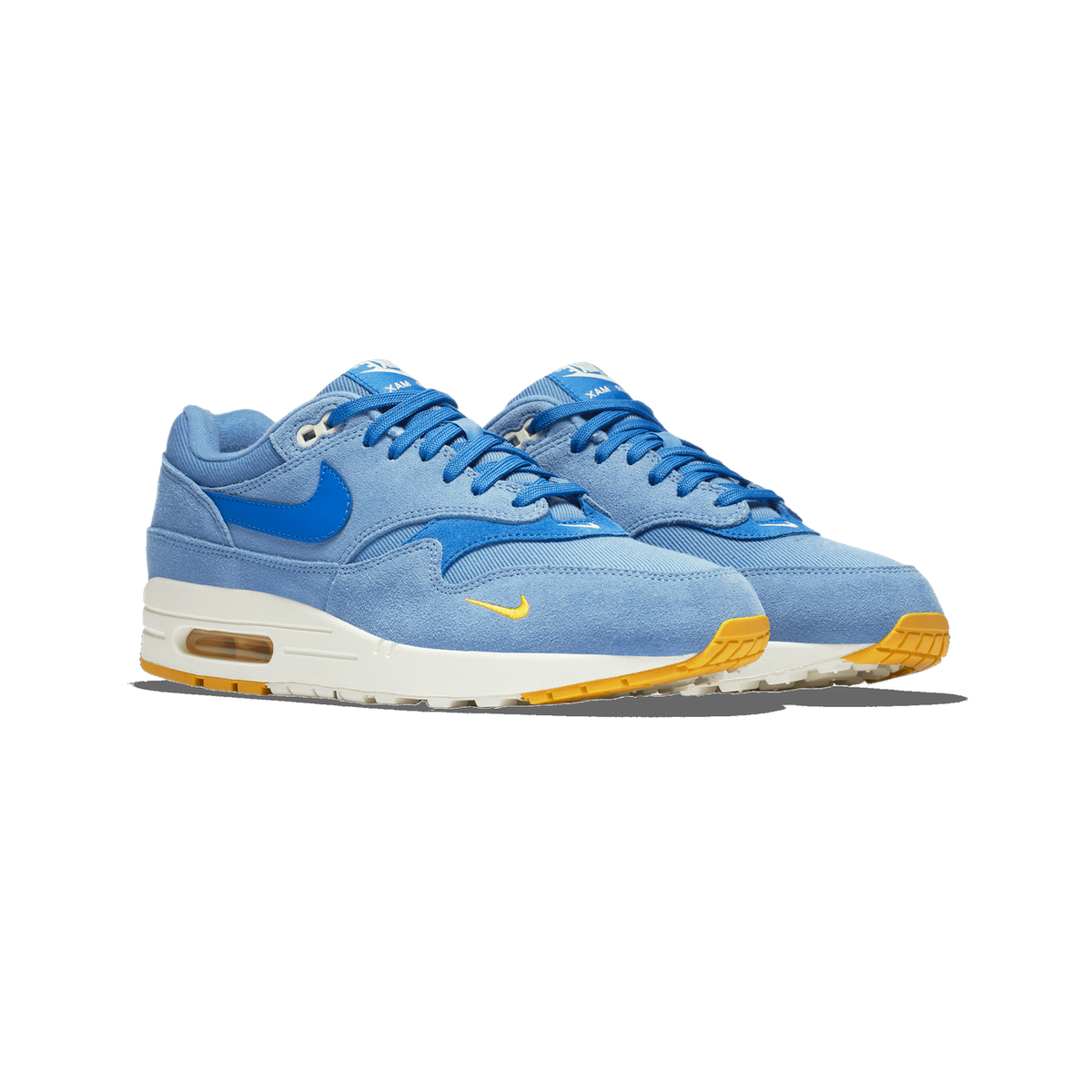 A pair of light blue suede Nike Air Max 1 sneakers shown side by side at an angle, with bright blue Swoosh logos, a small yellow Swoosh on the toe, blue laces, and yellow-orange outsole accents.