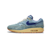Left Nike Air Max 1 sneaker in denim blue with a dark blue swoosh, cream laces, and a tan midsole with gum outsole, shown from the outside with toe pointing left.