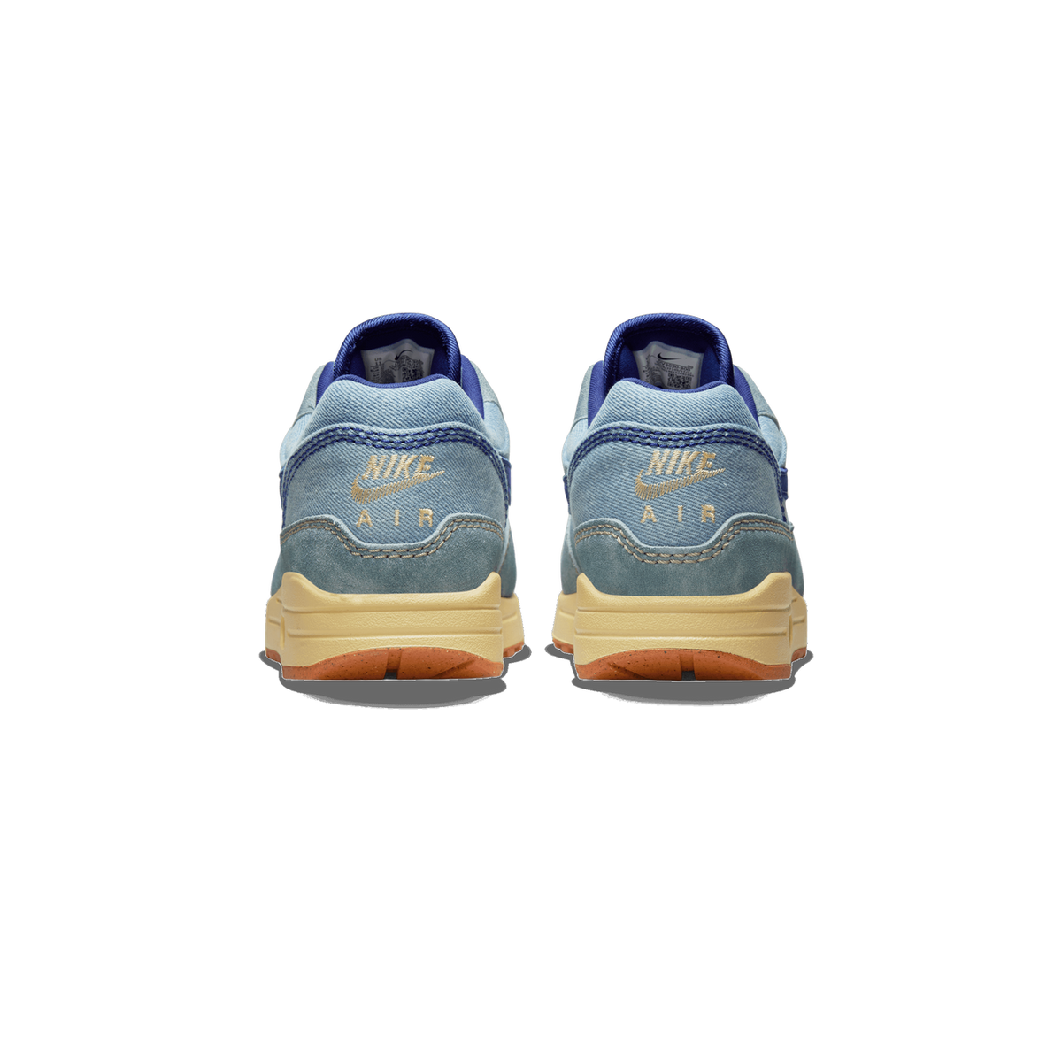 Rear view of a pair of blue denim Nike Air Max 1 sneakers with tan embroidered Nike Air logos and cream midsoles with gum outsoles.