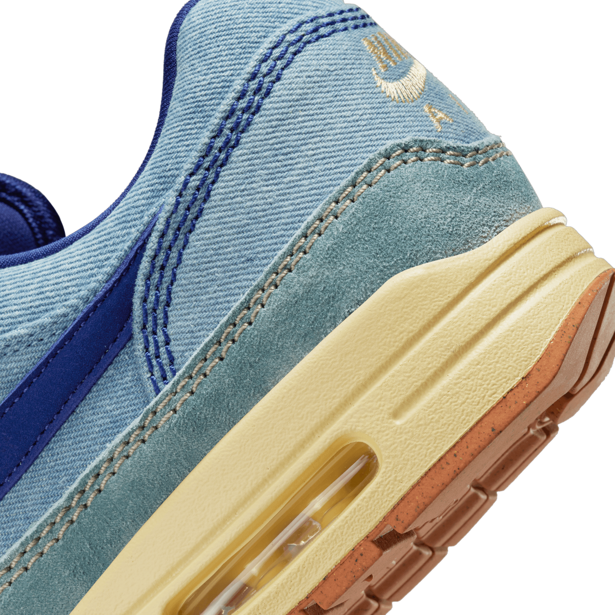 Close-up of the heel area of a blue denim Nike Air Max 1 showing the gold swoosh stitching, suede overlay, and visible Air Max cushioning unit in the cream midsole with gum outsole.
