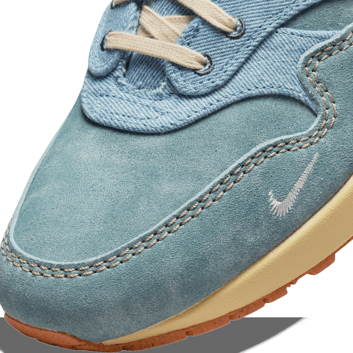 Close-up of the toe box of a blue suede and denim Nike Air Max 1 showing stitching detail, a small embroidered Swoosh, and a cream midsole with gum outsole