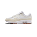 A single white and beige Nike Air Max 1 sneaker shown from the outside, toe pointing left, with white laces, a white Swoosh on the sail-coloured leather upper, beige suede toe and heel panels, a white midsole, and a visible clear Air unit with brown