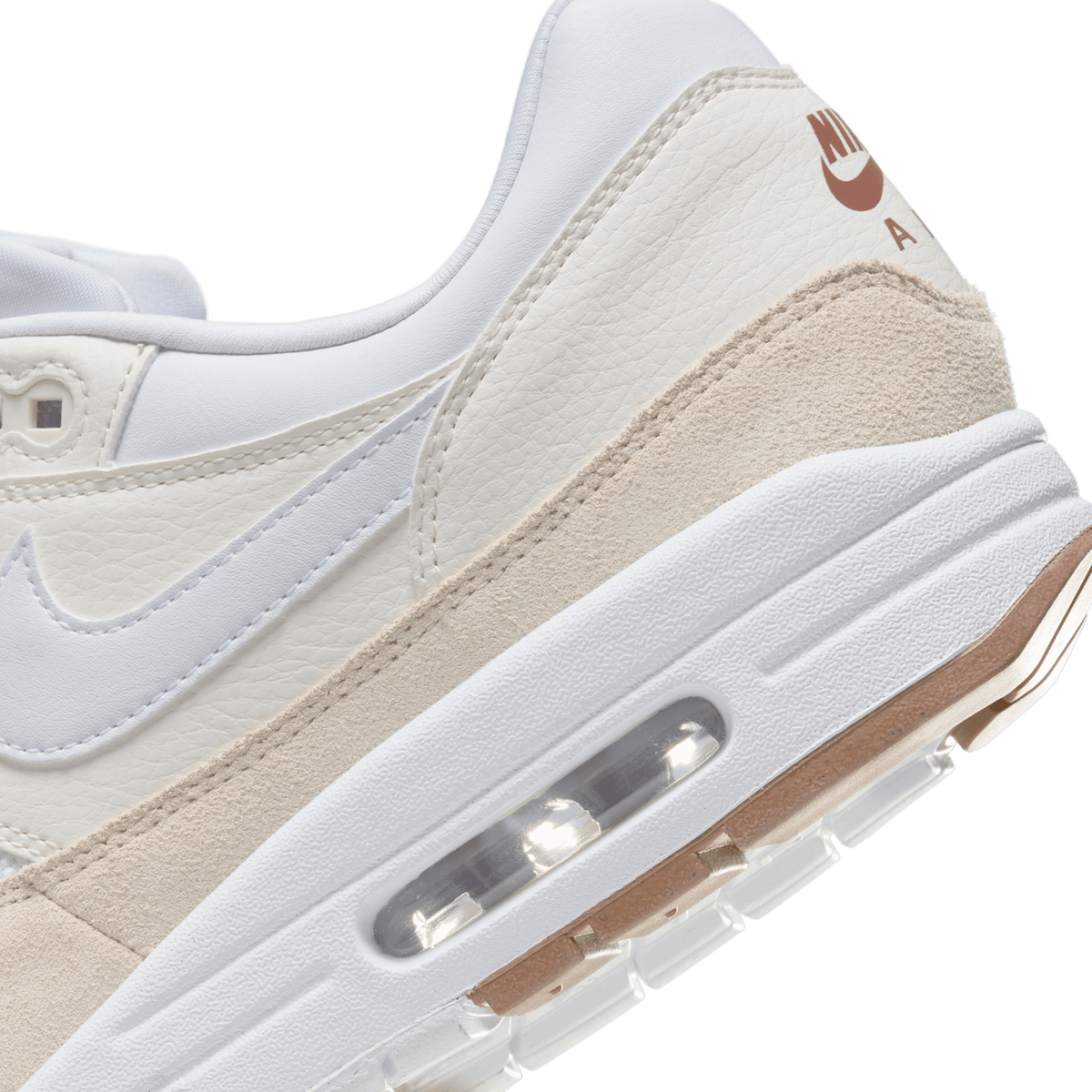 Close-up of the heel and midsole of a white and beige Nike Air Max 1 sneaker, showing the Nike logo and 'Air' lettering in brown on the heel tab, the beige suede panel, and the visible Air unit in the white midsole.