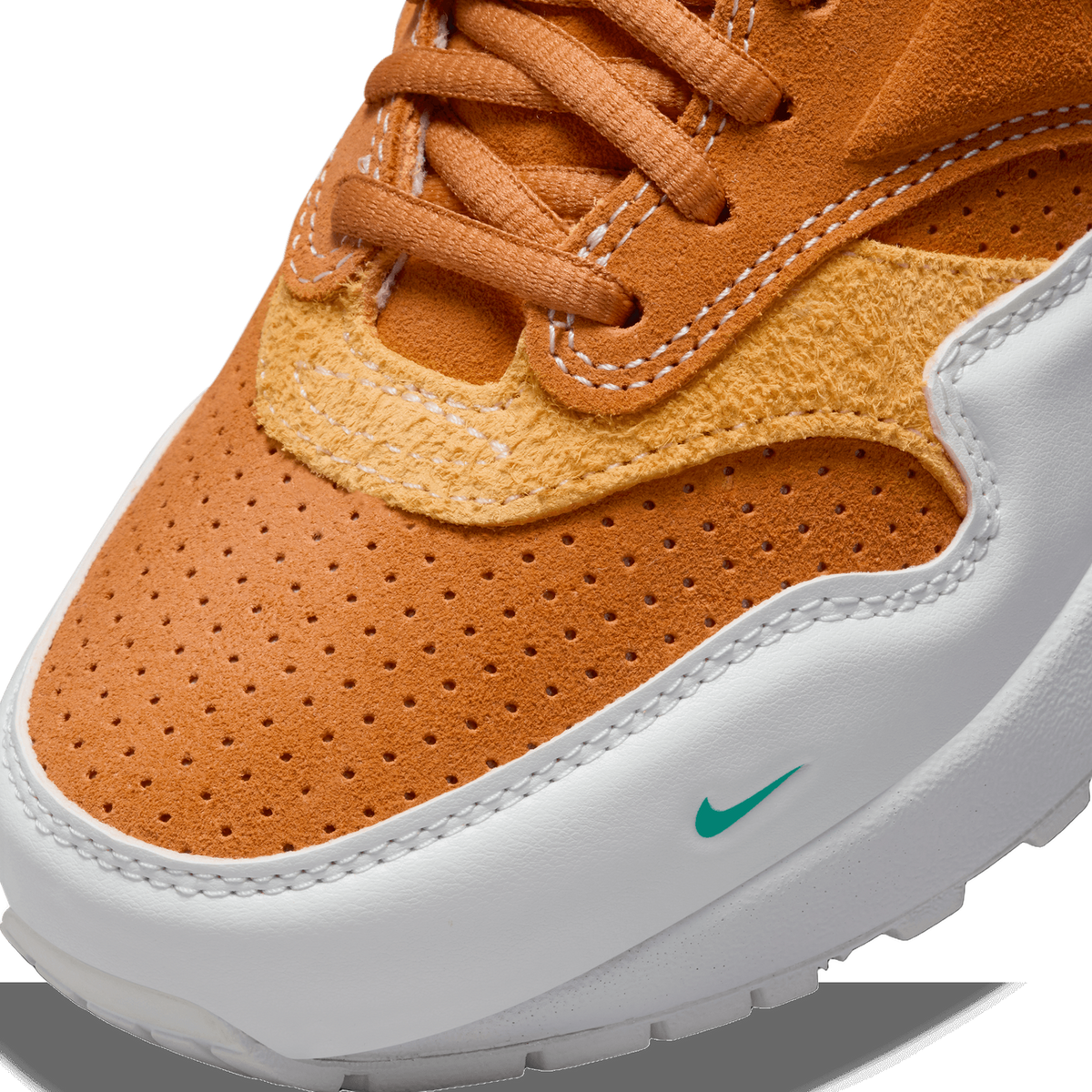 Close-up of a sneaker toe box with perforated burnt orange suede, tan textured overlay, white laces area, and a small green Nike swoosh on the white midsole panel