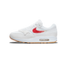 Side profile of a white Nike Air Max 1 with a red feather-style Swoosh embroidered on the side panel, white fur-like trim along the collar and tongue, grey suede toe and mudguard, and a visible clear Air unit in the white midsole with a gum rubber ou