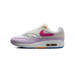 Left Nike Air Max 1 sneaker facing left, showing white and lavender panels, a pink swoosh, orange lace eyelets, and a visible Air unit with blue outsole accents