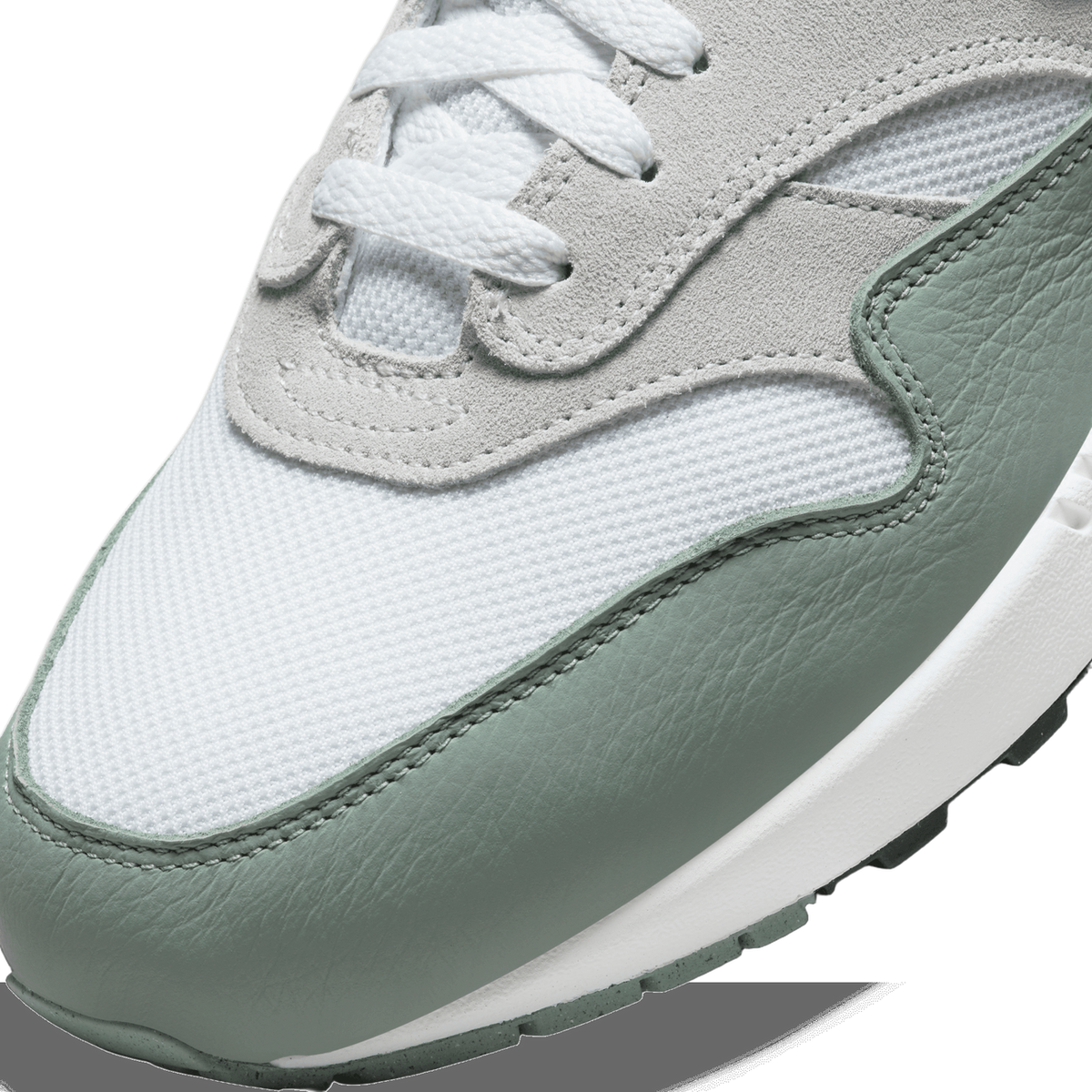 Close-up of a Nike Air Max 1 toe box showing white mesh, grey suede eyelet panel with laces, and green leather overlay above a white and green midsole