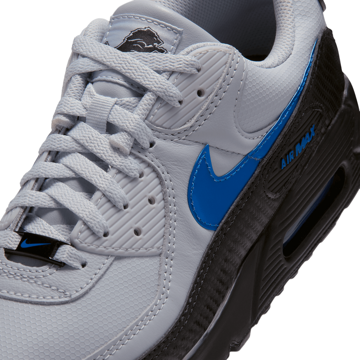 Close-up angled view of a grey Nike Air Max 90 showing the blue swoosh, laces, Lions logo tongue tag, and black Air Max midsole detailing.