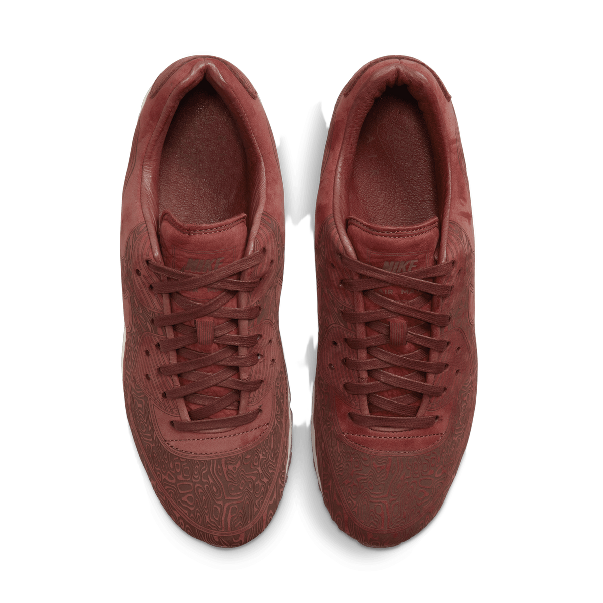 Overhead top-down view of a pair of rust-brown Nike Air Max 90 sneakers placed side by side, showing laser-etched topographic patterns across the toe and overlays, laces, and the tongues with a visible NIKE logo.