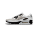 Lateral view of a white and grey Nike Air Max 90 with black heel overlay, black lace panel, orange 'Air Max' branding, and visible heel air unit, toe pointing left.