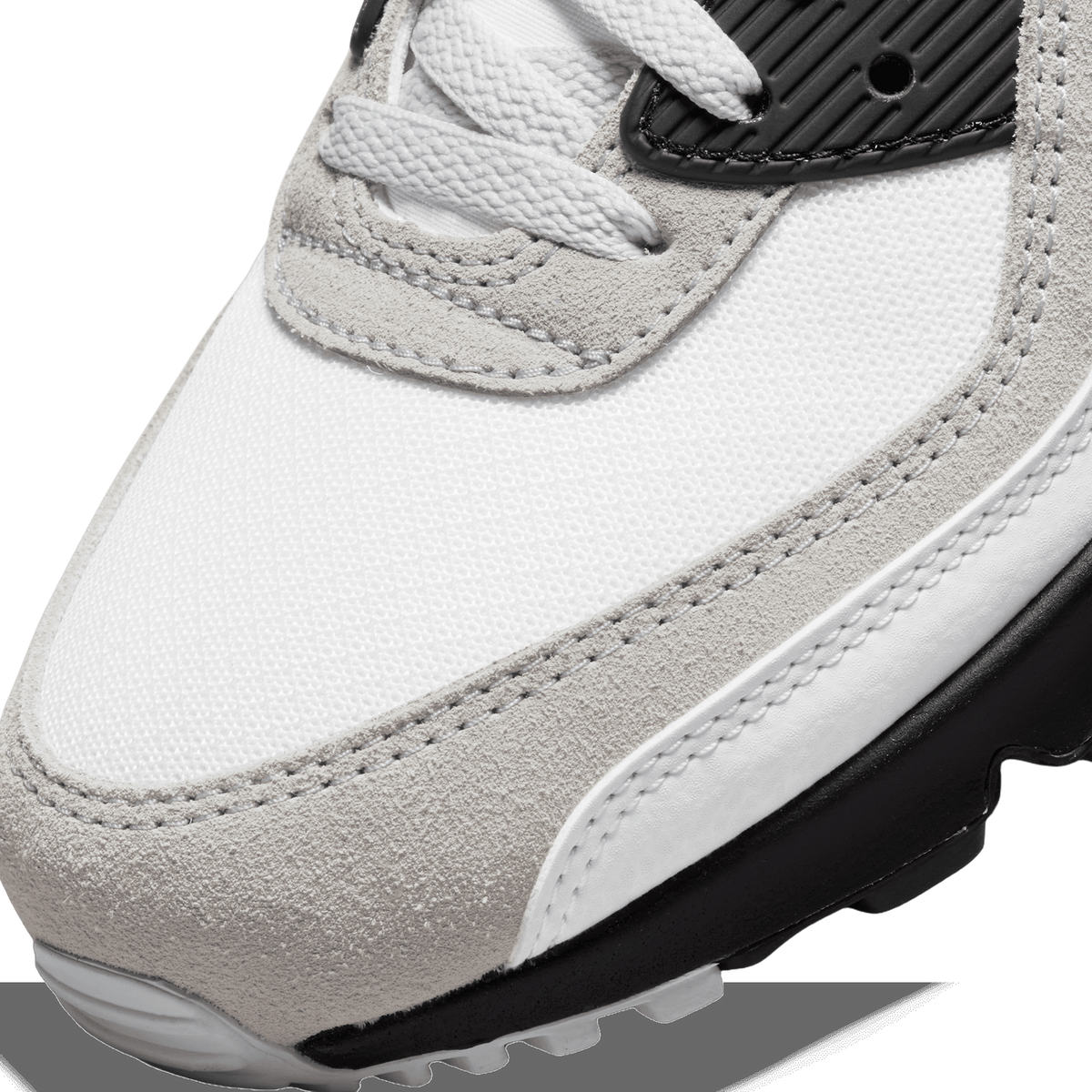 Close-up of the toe box of a white and grey Nike Air Max 90 sneaker with suede overlays, mesh panels, and a black heel panel visible in the background.
