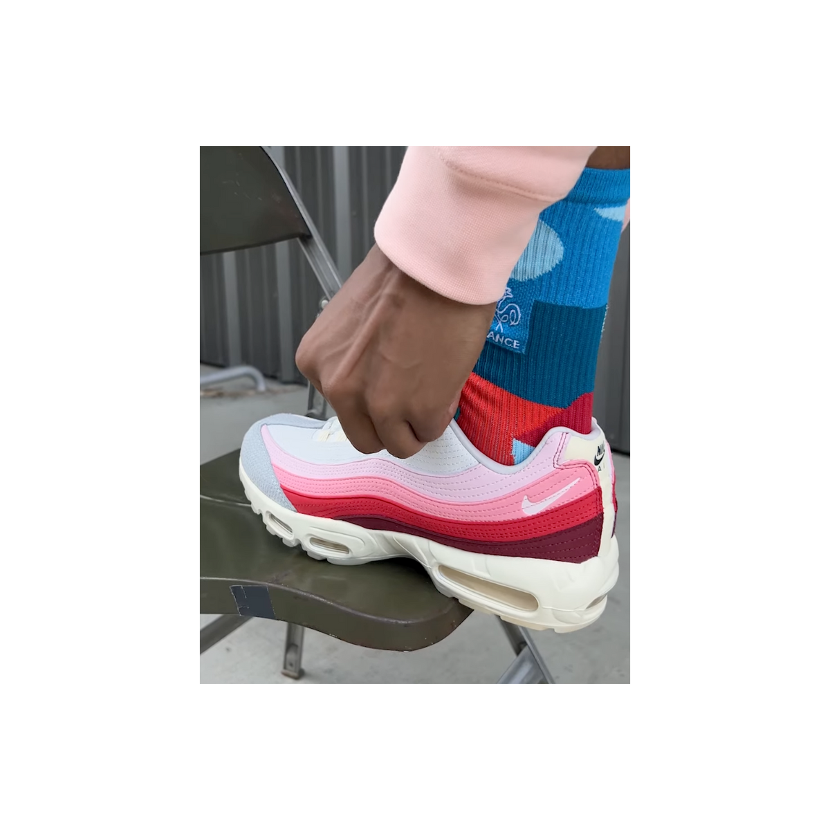 A person wearing a pink, red, and grey Nike Air Max 95 with layered gradient panels and a visible Swoosh logo, foot resting on a folding chair.