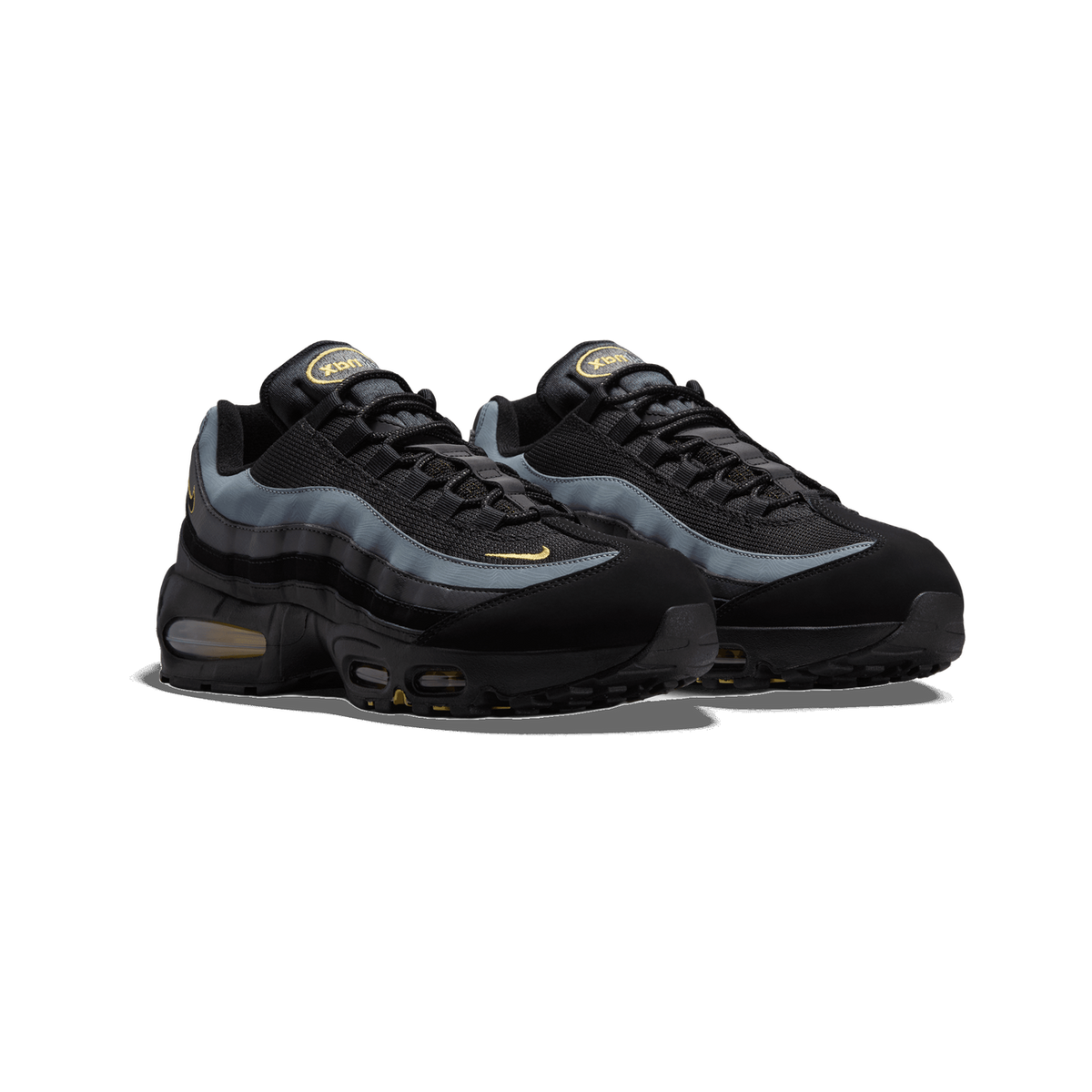 A pair of black and dark grey Nike Air Max 95 sneakers angled side by side, showing the grey mesh panels, black suede-like overlays, yellow Swoosh logos, and visible yellow Air units in the midsole.