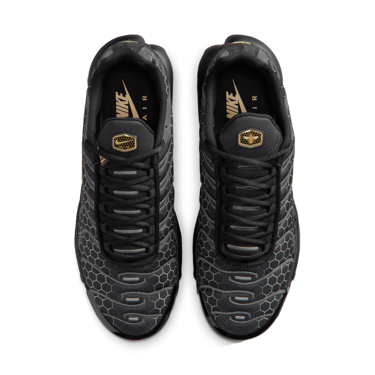 Top-down view of a pair of black Nike Air Max Plus sneakers with honeycomb textured uppers and gold branding on the insoles and heel tabs