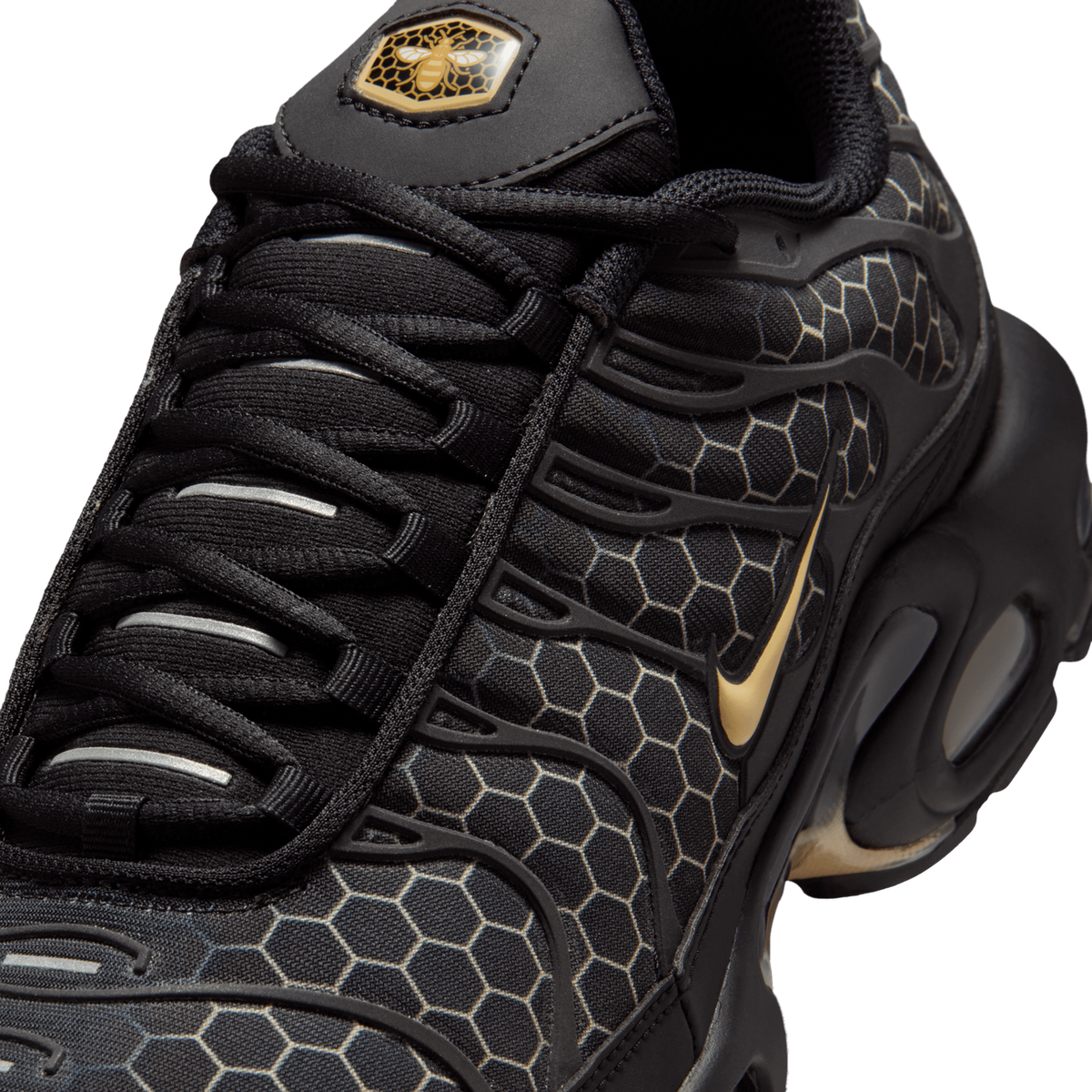 Close-up of a black Nike Air Max Plus with honeycomb pattern overlay, gold Swoosh logo, and a bee-and-honeycomb badge on the heel tab.