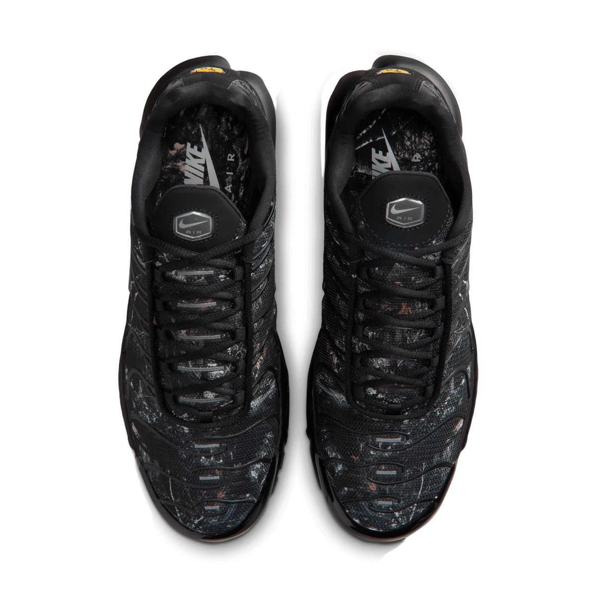 Top-down view of a pair of black Nike Air Max Plus sneakers with a wood camo print pattern and branded pull tabs on the tongues.