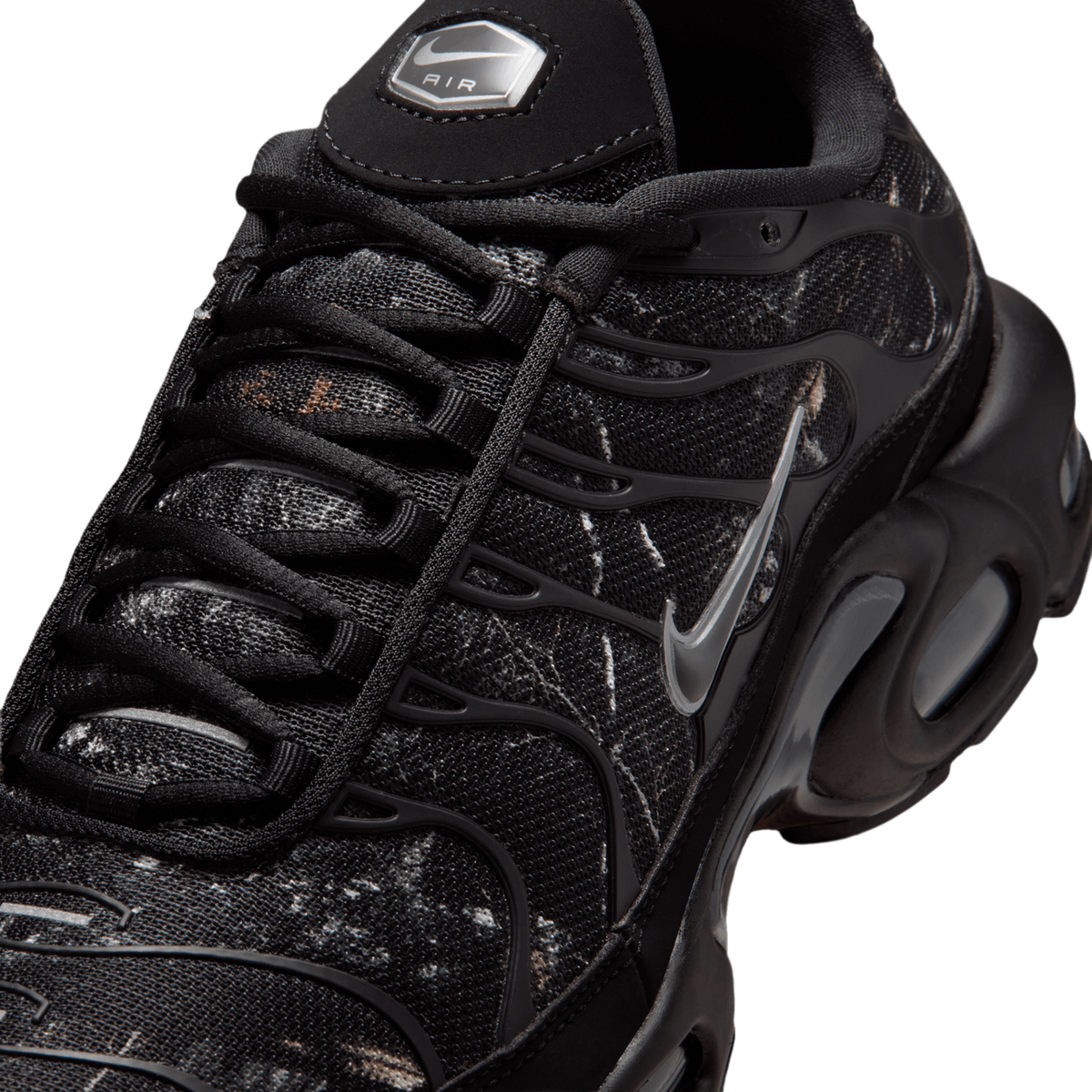 Close-up top view of a black Nike Air Max Plus sneaker with camo-textured overlays, wavy TPU panels, a silver Swoosh, and an Air logo tab on the tongue.