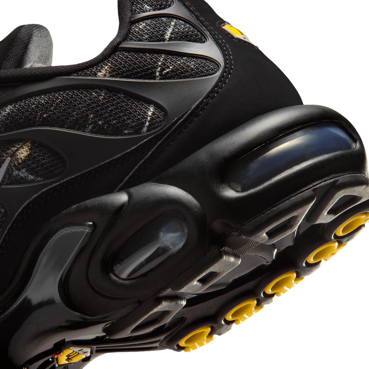 Close-up angled view of a black Nike Air Max Plus sneaker showing the heel Air unit, textured upper panels, and the yellow-accented outsole tread.