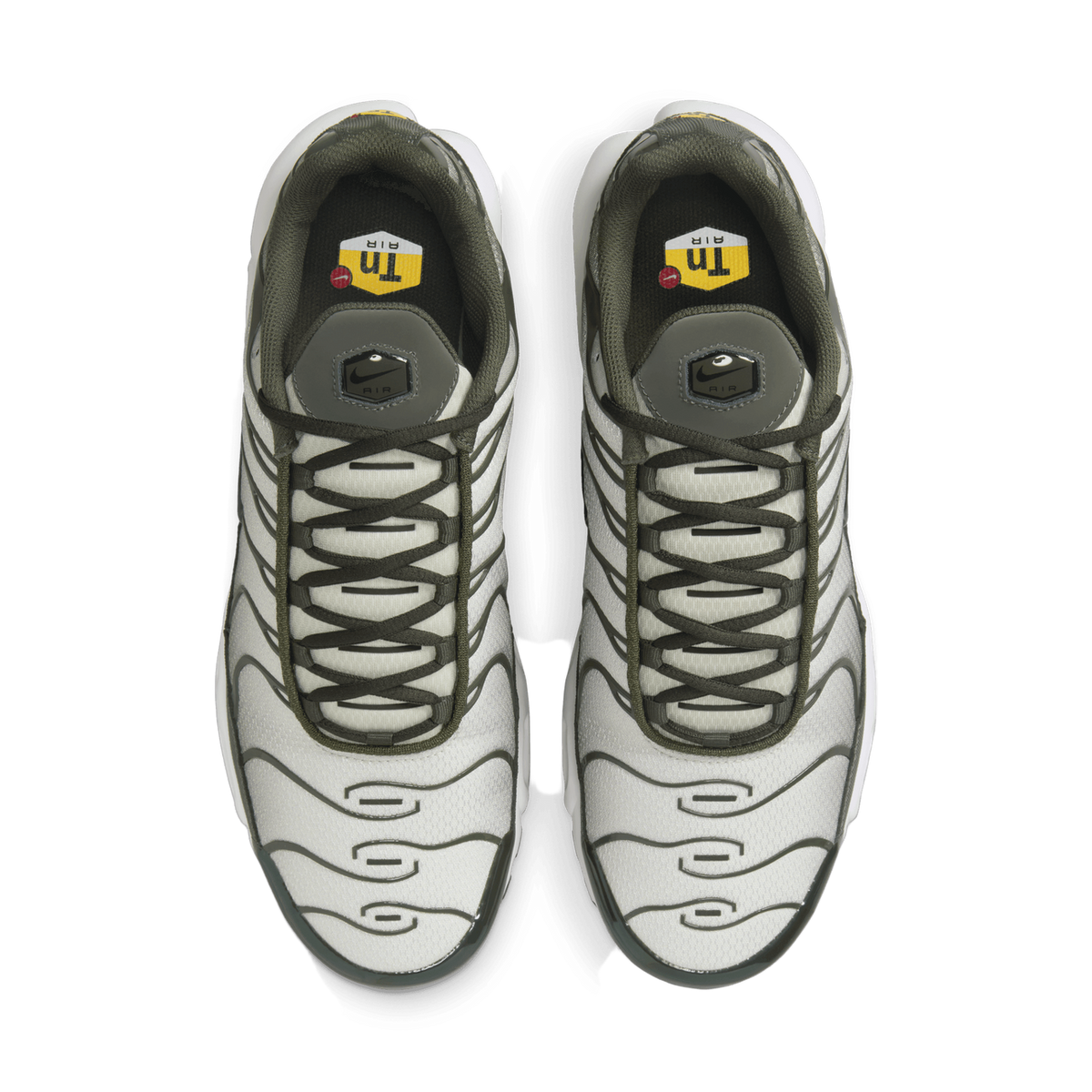 Top-down view of a pair of Nike Air Max Plus sneakers with light grey mesh uppers, dark olive green overlays and laces, and a visible yellow and white 'Tn Air' tongue label inside the collars, placed side by side with toes pointing toward the viewer.