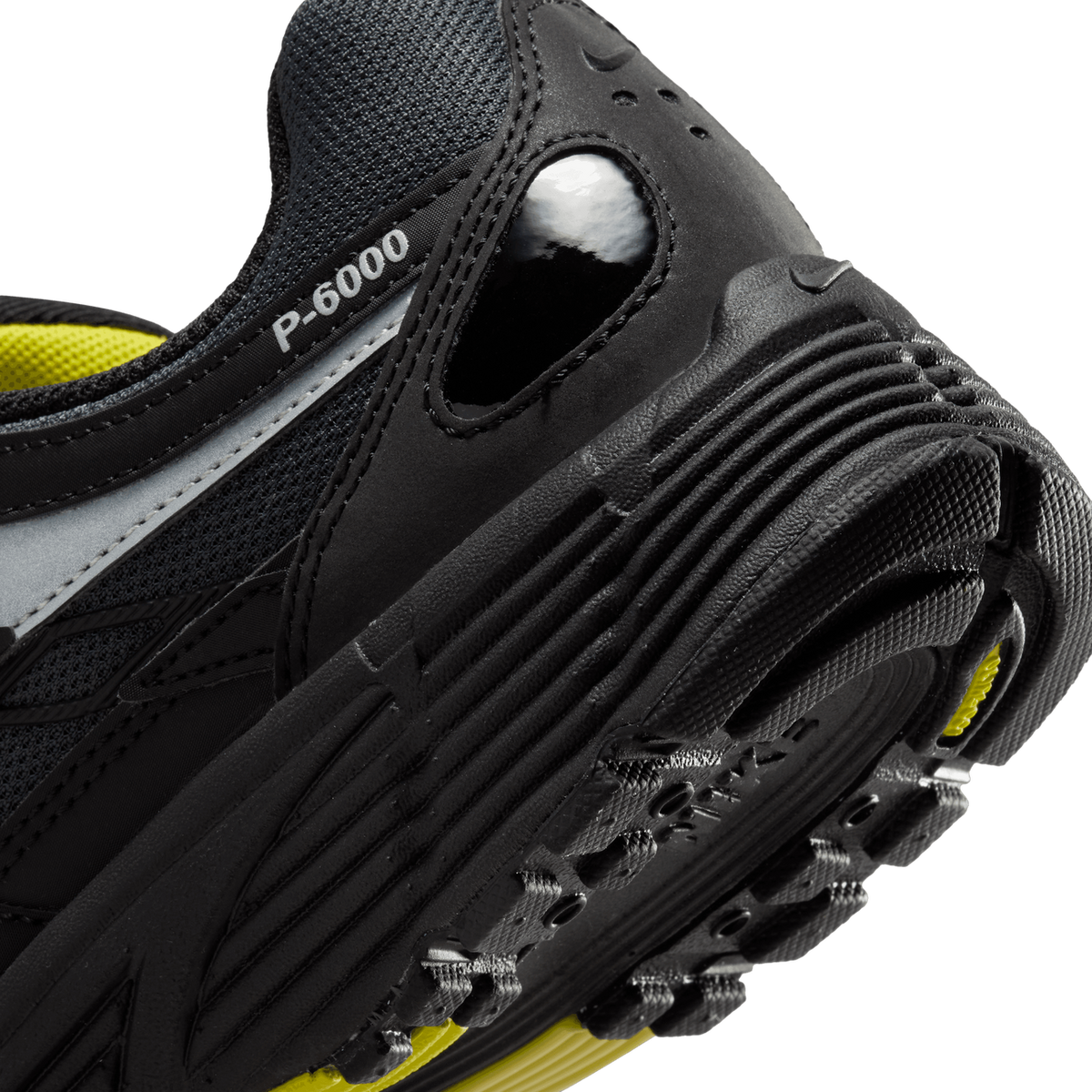 Close-up of the heel of a black Nike P-6000 sneaker showing the 'P-6000' text on the side panel, a grey reflective overlay, a glossy black heel tab with a pull loop, and a ridged black outsole edge with a glimpse of yellow midsole.