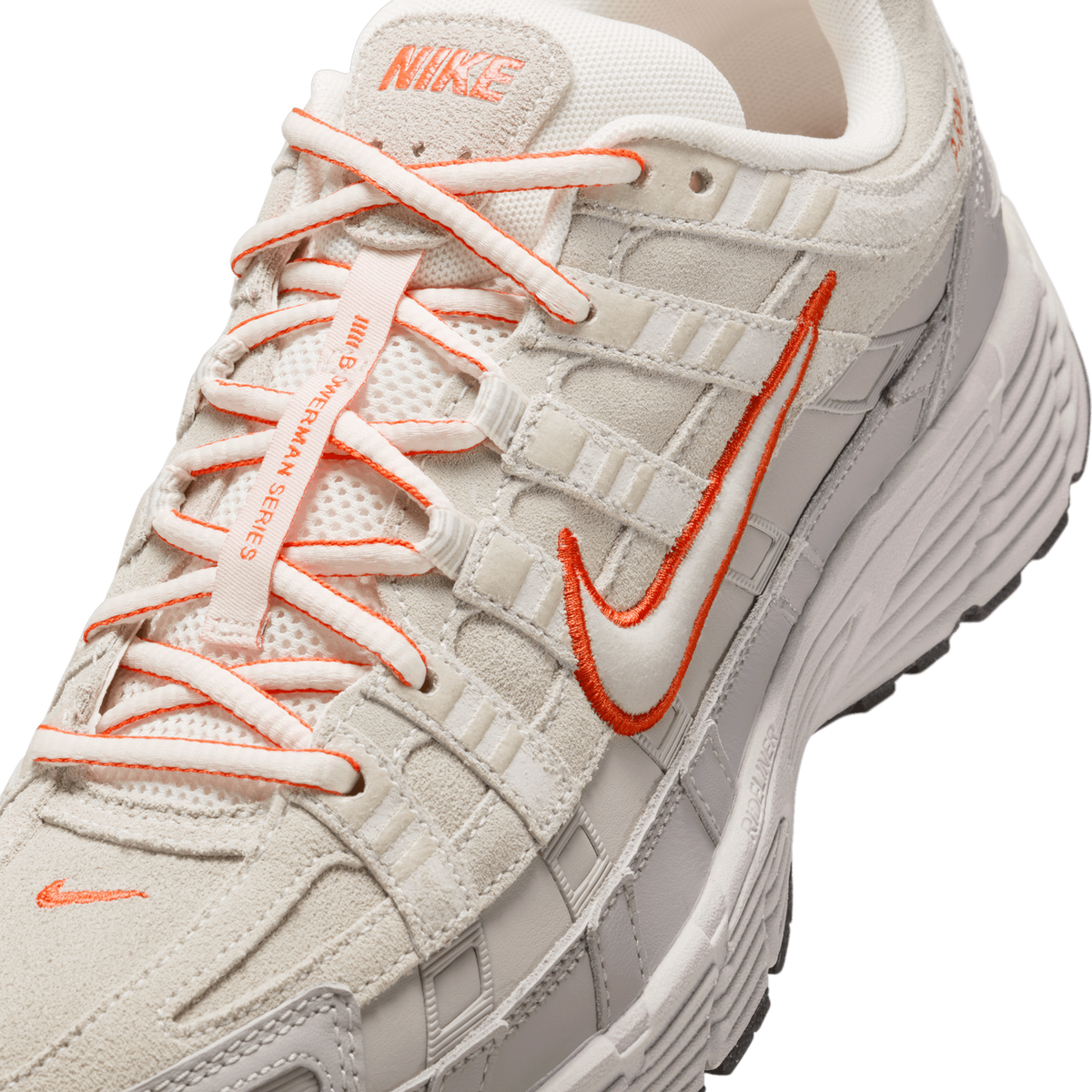 Close-up detail of a cream and light gray Nike sneaker showing the tongue with a "NIKE" label, orange-accented laces with a Ski Mask-style lace tag, and an orange embroidered Swoosh on the side panel.