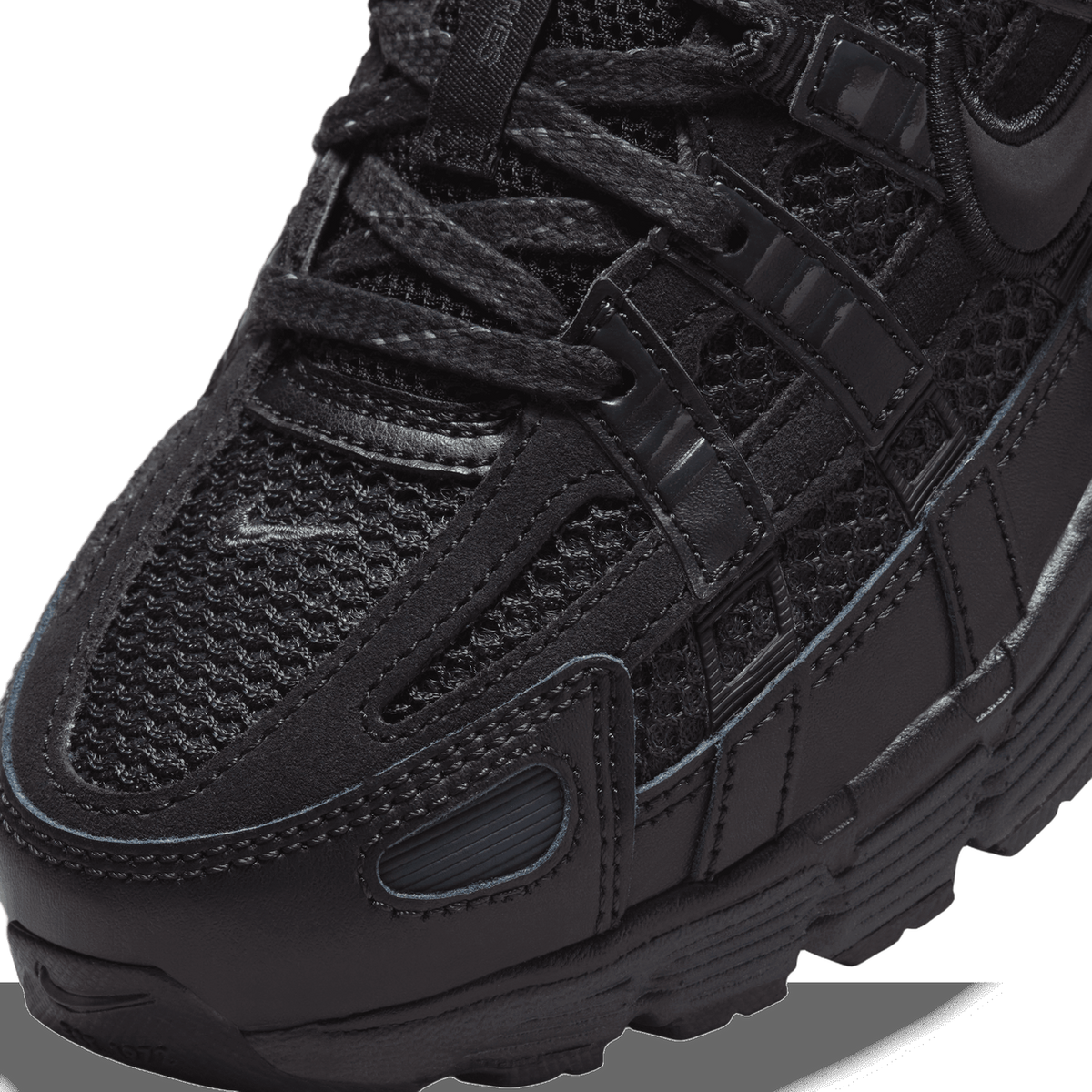 Extreme close-up of a black Nike P-6000 sneaker showing the textured black mesh panels, black suede overlays, glossy black Nike swoosh tabs, and the lace-up area with flat black laces.
