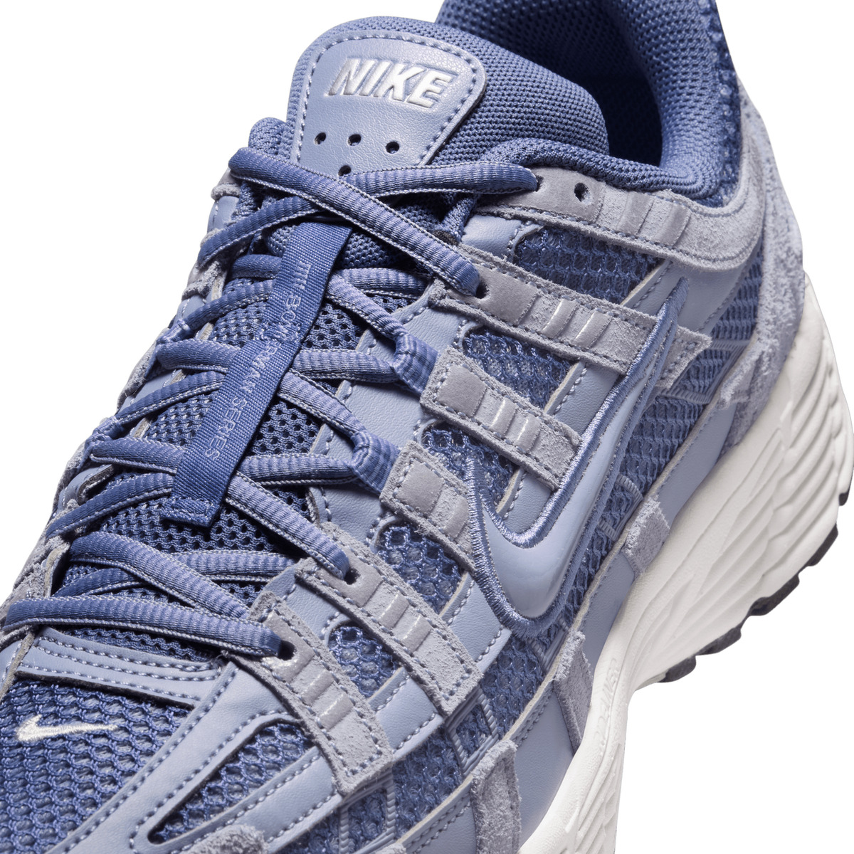 Close-up detail of a blue and grey Nike P-6000 sneaker showing the laces, heel tab with embossed NIKE logo, mesh and suede panels, and the Swoosh stitched on the side with a white midsole.