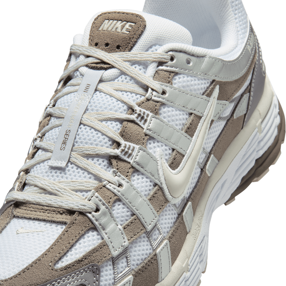 Close-up top view of a white Nike P-6000 sneaker with brown suede overlays, a cream swoosh, and laces with a webbing pull tab reading 'Roman Series'