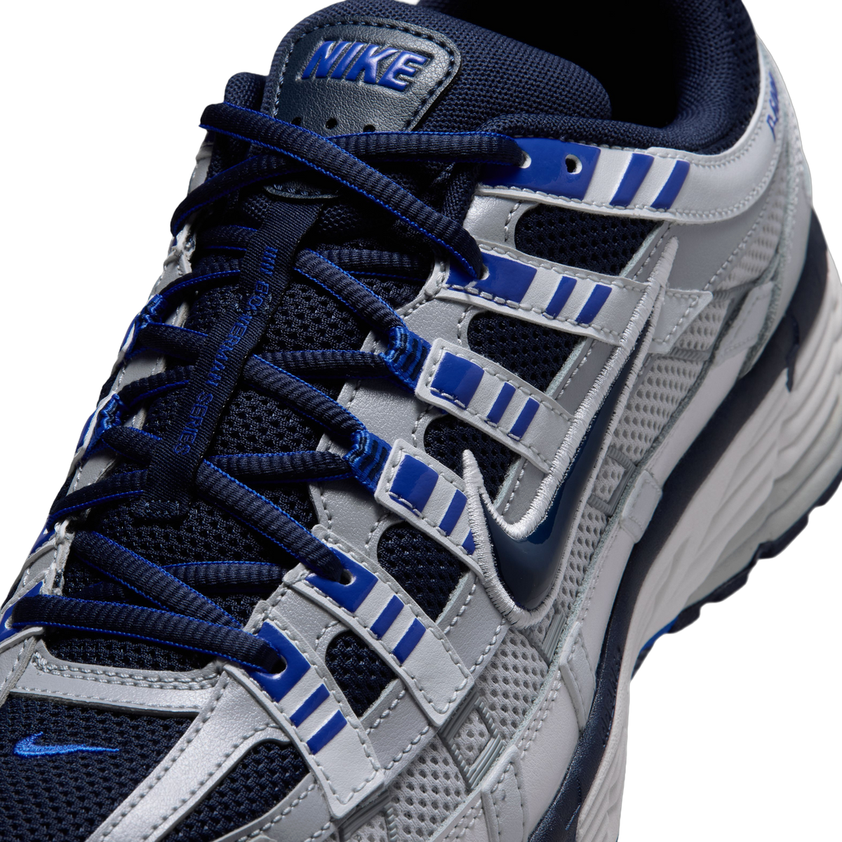 Close-up of a Nike P-6000 sneaker's lace eyelets, tongue, and swoosh in gray, navy, and blue with visible Nike branding.