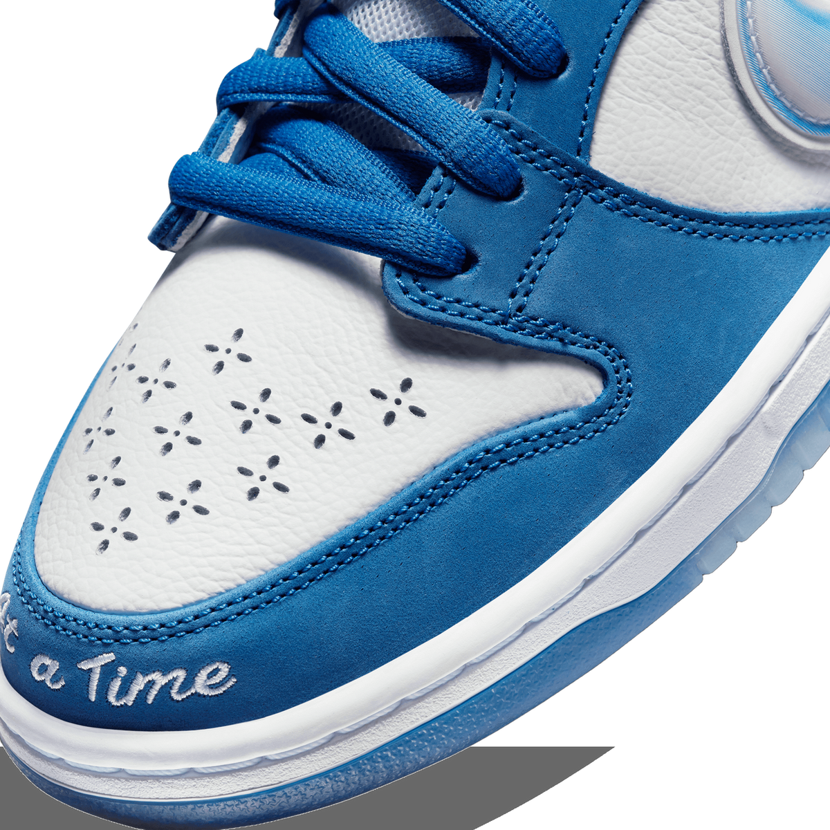 Close-up of the toe box of a blue and white Nike SB Dunk Low showing perforated flower pattern detailing and 'a Time' embroidered text near the sole
