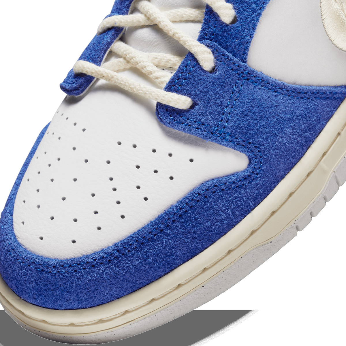 Close-up of a sneaker toe box showing blue suede overlay, white leather perforated panel, and white laces threaded through blue eyelets.