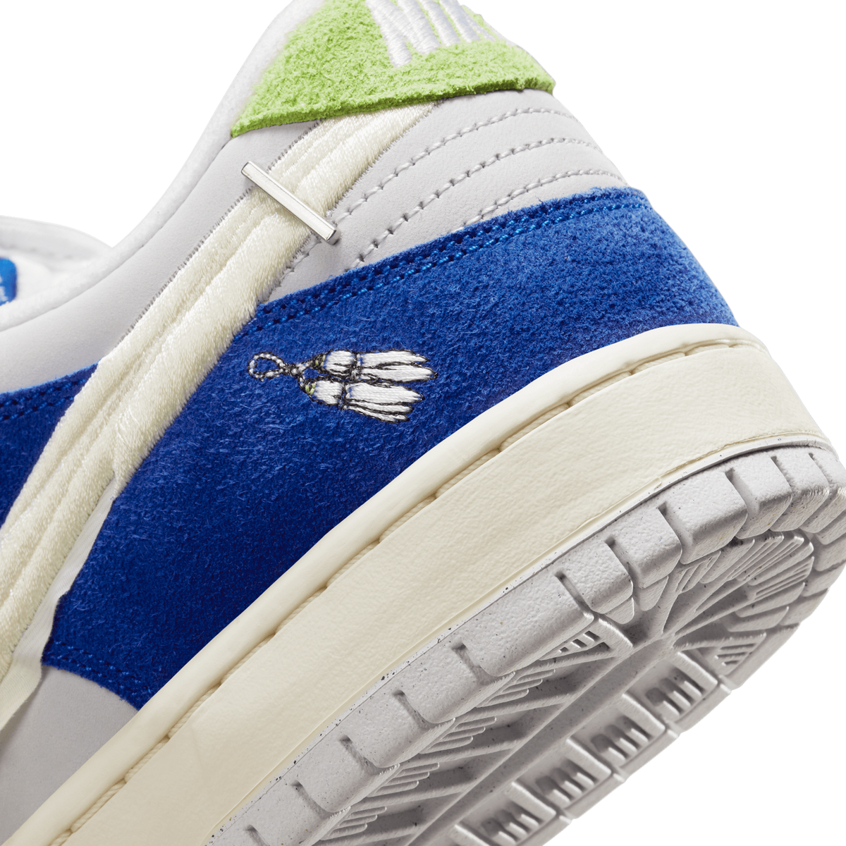 Close-up angled view of a Nike SB Dunk Low showing blue suede panel with embroidered flower design, cream laces, green suede heel tab, and textured outsole.