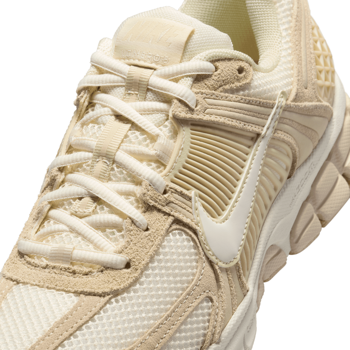 Close-up detail of a cream and beige sneaker showing the laced tongue with a tan Nike Vomero label, white laces, tan suede overlays, mesh panels, and a white Swoosh on the side.